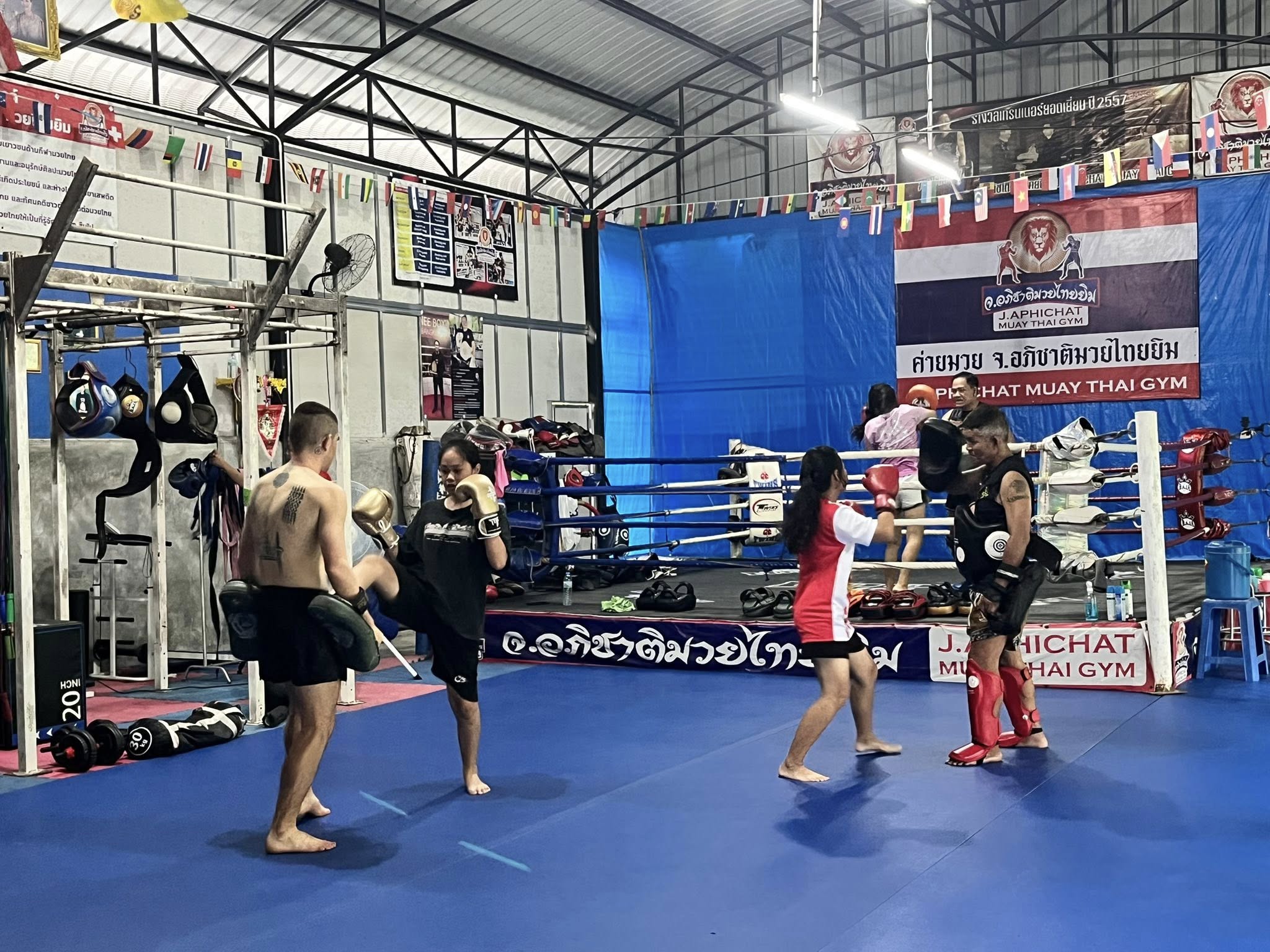 Students training together inside J. Aphichat Muay Thai Gym, with a boxing ring and the camp's banner in the background, Sing Buri