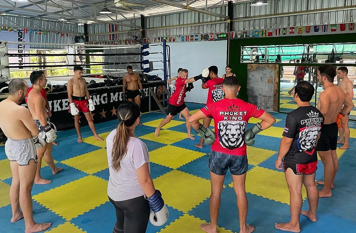 Muay Thai coach demonstrating striking techniques to a group class at Phuket King Muay Thai, as students gather in a circle to watch and learn in a lively, hands-on training session.