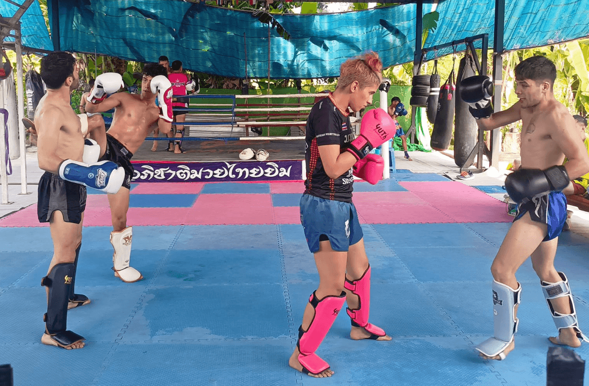 A group of Thai and international students trainig together in front of the J. Aphichat Muay Thai Gym banner, Sing Buri
