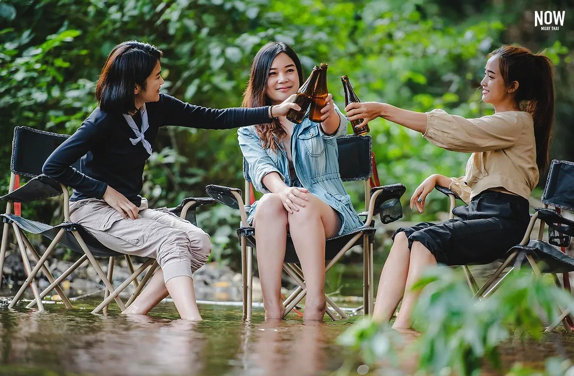 At Mae Kampong, a traditional village in Chiang Mai surrounded by lush nature, three female travelers relax and enjoy beers while sitting in a cool, gentle stream that flows through the village’s serene setting.