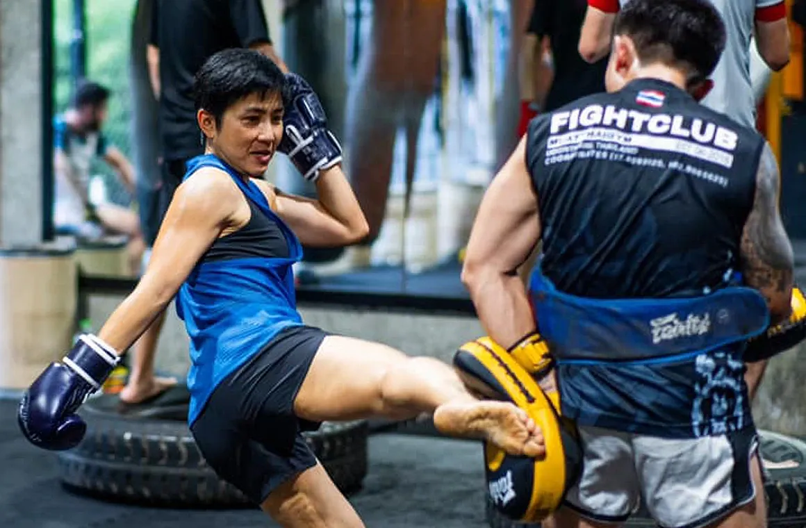 A female fighter practices a roundhouse kick during a Muay Thai group class for fighters at Fight Club Muay Thai Gym, with Kru Parker providing feedback.