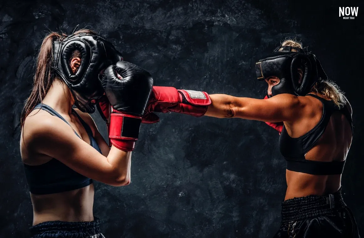 Two female kickboxers in full protective gear, including headguards and gloves, sparring intensely in a training ring.