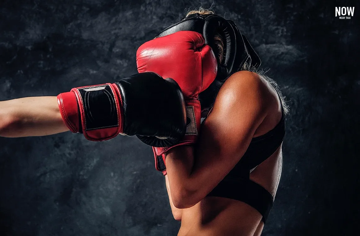 An intense close-up shot captures a female kickboxer mid-fight, she is protecting her head with the gloves while another kickboxer tries to punch her in the face