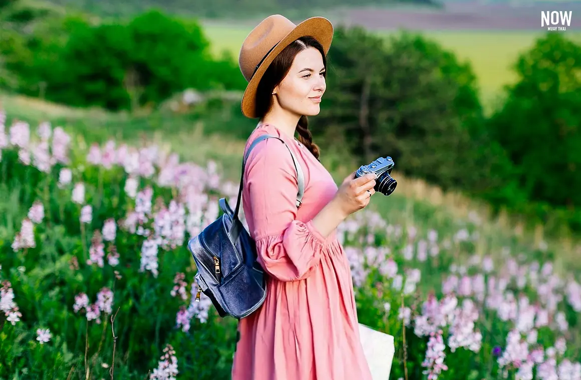 A woman with a hat, a denim backpack and a pink dress, holding a camera and a map, standing  in a flower field at Singha Park, Chiang Rai, Thailand, sprawling flower fields, vibrant blooms, picturesque landscape, winter season