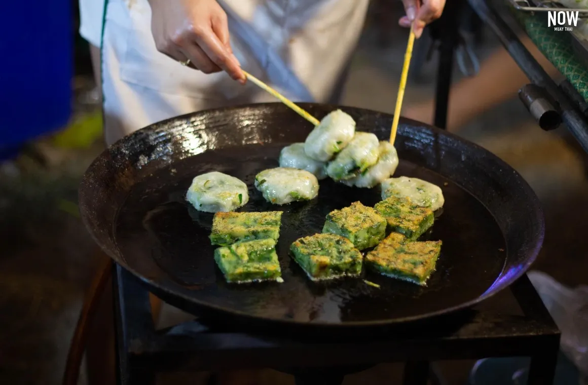 Close-up shot of a frying pan filled with Chinese chive dumplings (kui chai), some appearing freshly fried and others being stir-fried with a dark sauce, at a busy Bangkok street food stall.