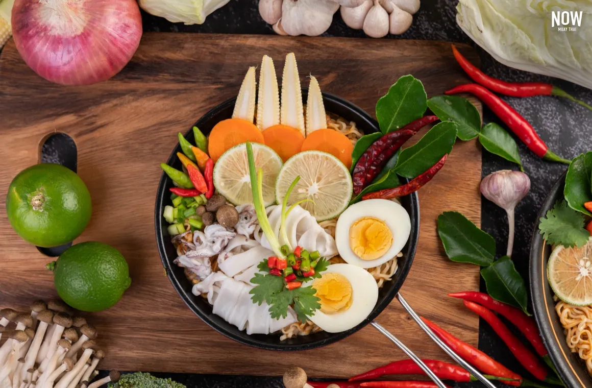 A close-up shot of a steaming bowl of Tom Yum Mama (instant noodles in spicy tom yum broth) topped with large slices of squid, half a boiled egg, and fresh herbs, sitting on a table.