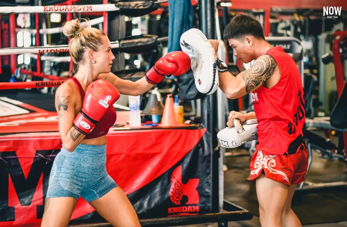 a blonde woman hitting a trainer’s glove with her left hand, practising muay thai techniques