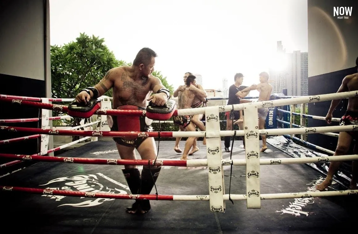 a man standing on the edge of a boxing ring at Elite Fight Club Bangkok, looking behind himself he sees 4 people practicing muay thai fight and in the background we can see some skyscrapers of Bangkok
