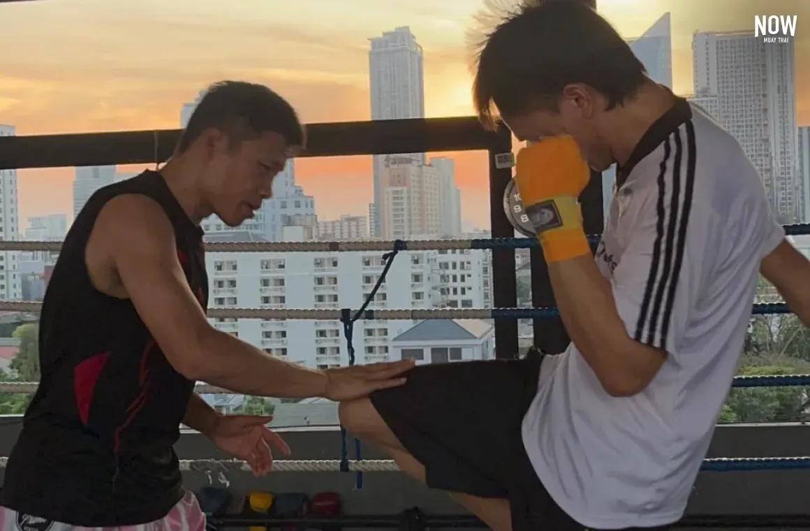 2 men practicing muay thai kicks with knee at Tded99 Muay Thai Gym in Bangkok, behind them we see the sunset and high skyscrapers of Bangkok