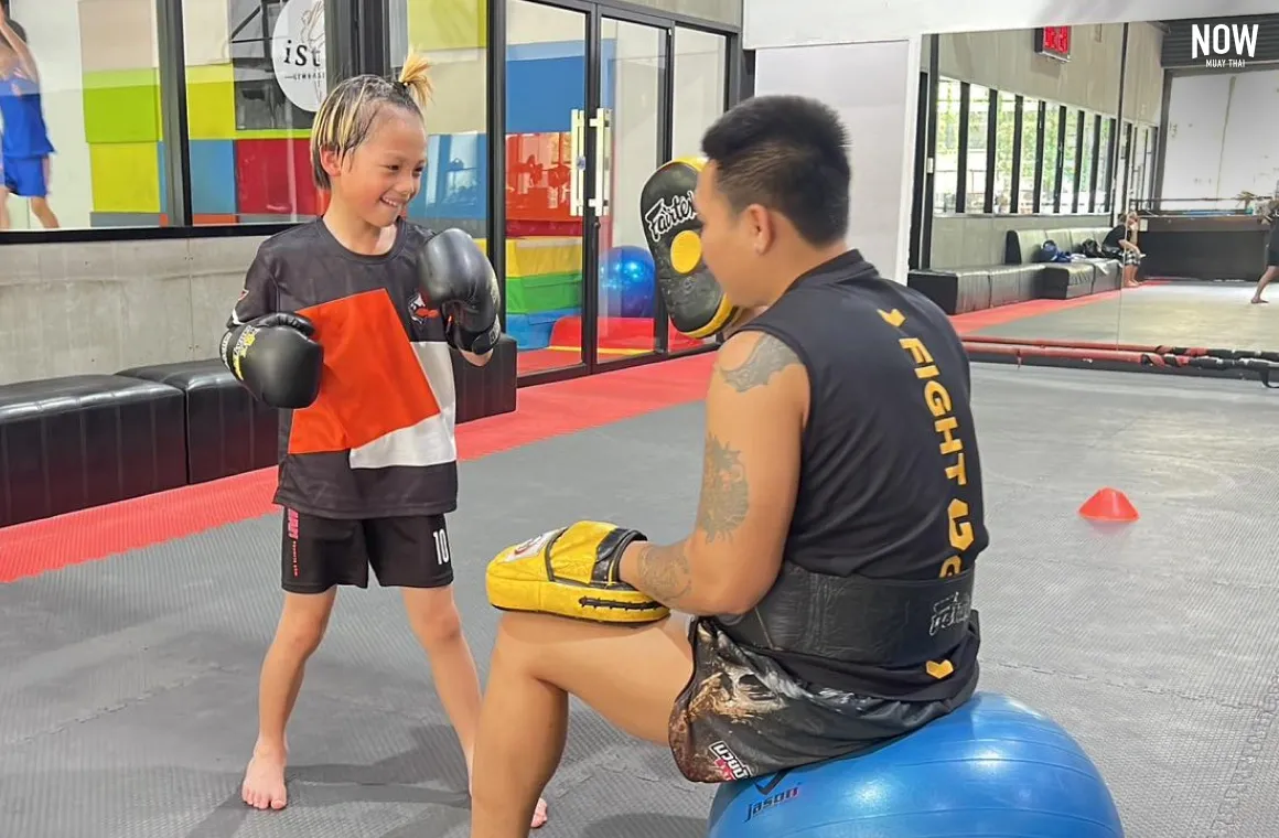 trainer sitting on a blue fitness ball awaiting a punch in the glove from a 6 year old kid, that looks very happy and excited to try muay thai at 7 Stars Muay Thai Gym