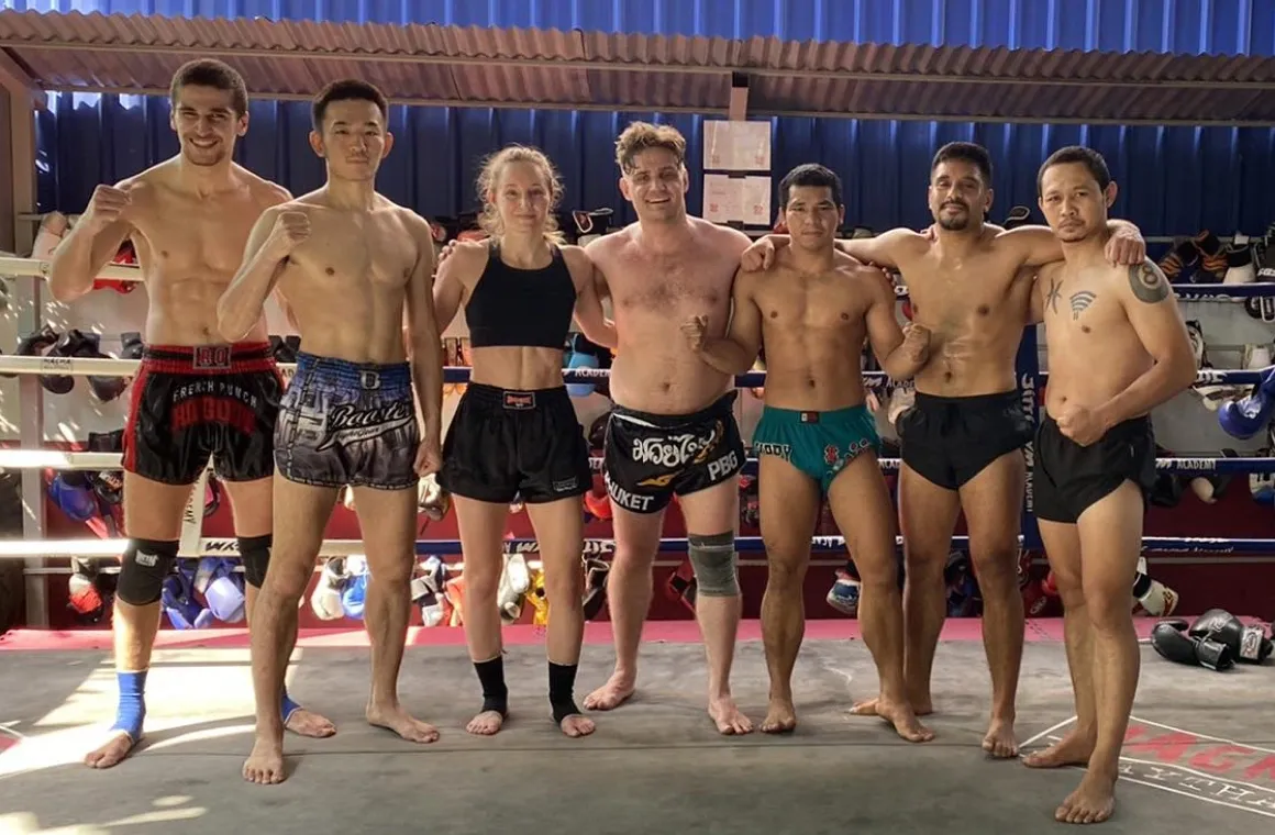 7 people taking a group picture, standing in front of the boxing ring at Rithirit Gym Academy in Bangkok. There are 6 men and one woman all dressed with muay thai shorts