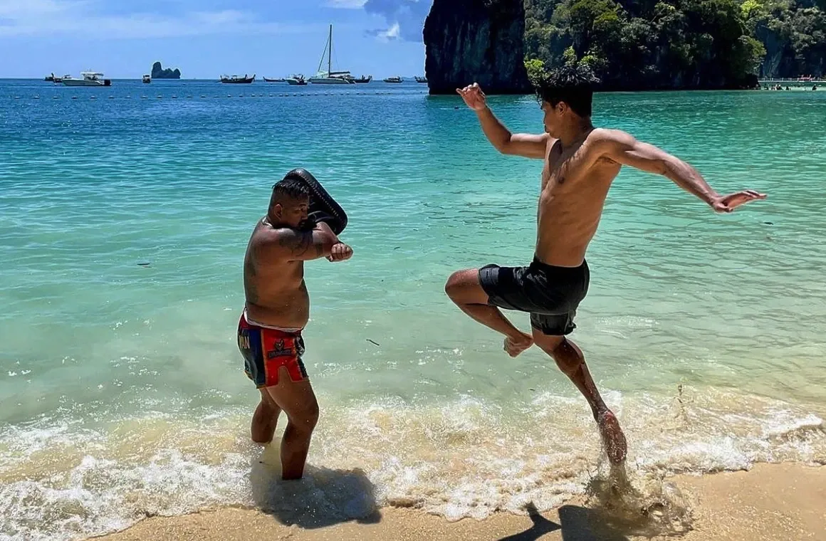 Muay Thai champion Superbon practicing with his trainer - Trainer Gae on a sandy beach in Thailand, captured mid-air as he performs a powerful jump kick toward his trainer amidst a tropical beach setting.
