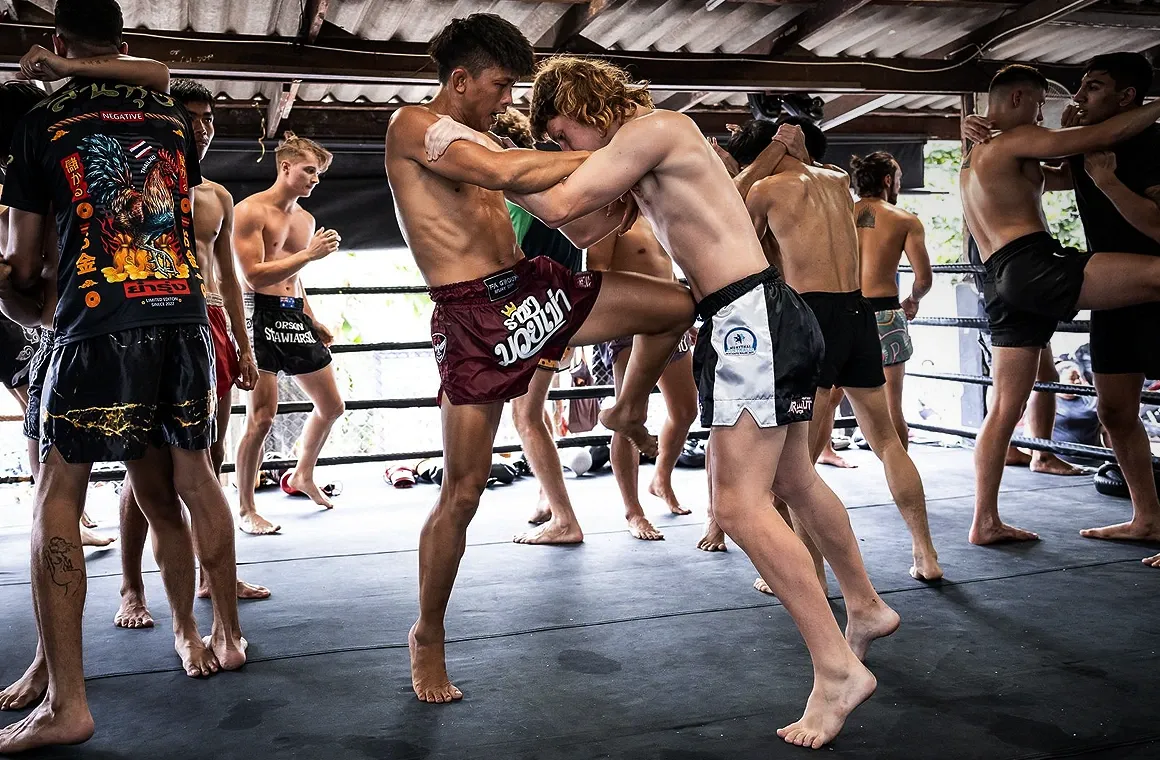 Authentic Muay Thai training session led by Kru Yothin FA Group, a renowned trainer with titles including 2x National Thailand Champion, RWS Champion, WBC International Champion, and Rajadamnern Stadium Champion. The image captures Kru Yothin guiding students through traditional techniques, highlighting the dedication and skill in an authentic Muay Thai gym setting.