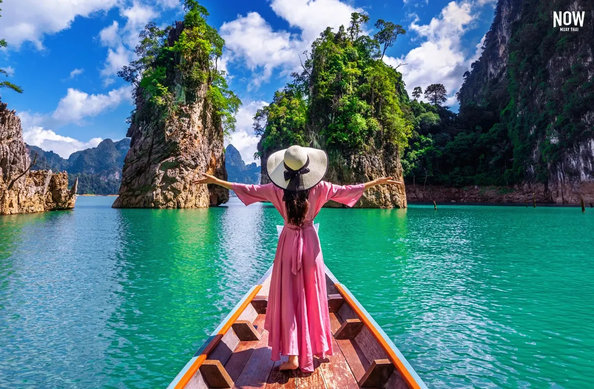 Female tourist standing on a small boat, breathing fresh air surrounded by lush green mountains in Thailand – Thailand is more than a place to visit; it’s a destination to recharge with yourself