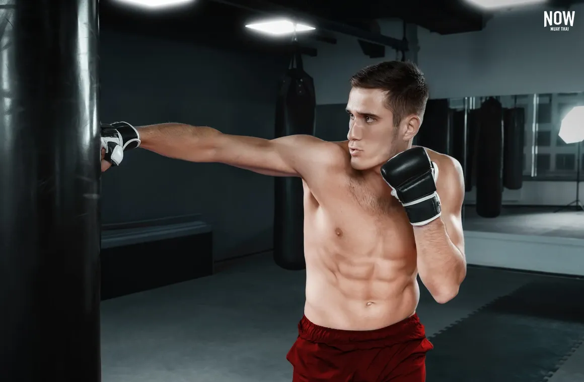 Young male Muay Thai fighter in black gloves practicing hand punches on a punching bag in the gym, demonstrating how Muay Thai builds strength beyond traditional muscle gains.