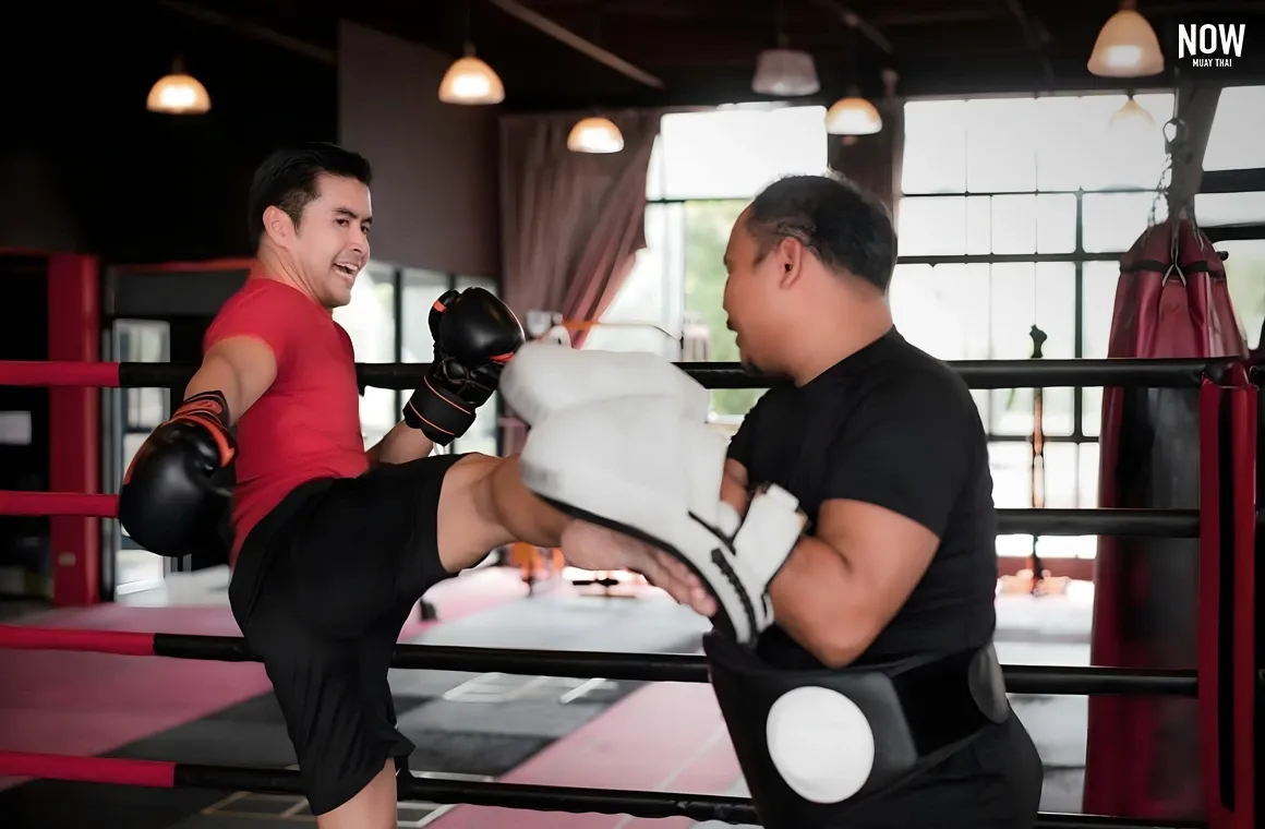 A male student having fun as he practices kicking a target held by his Muay Thai trainer. The instructor provides hands-on support, guiding the student through proper technique and offering encouragement, ensuring a productive and enjoyable training session.
