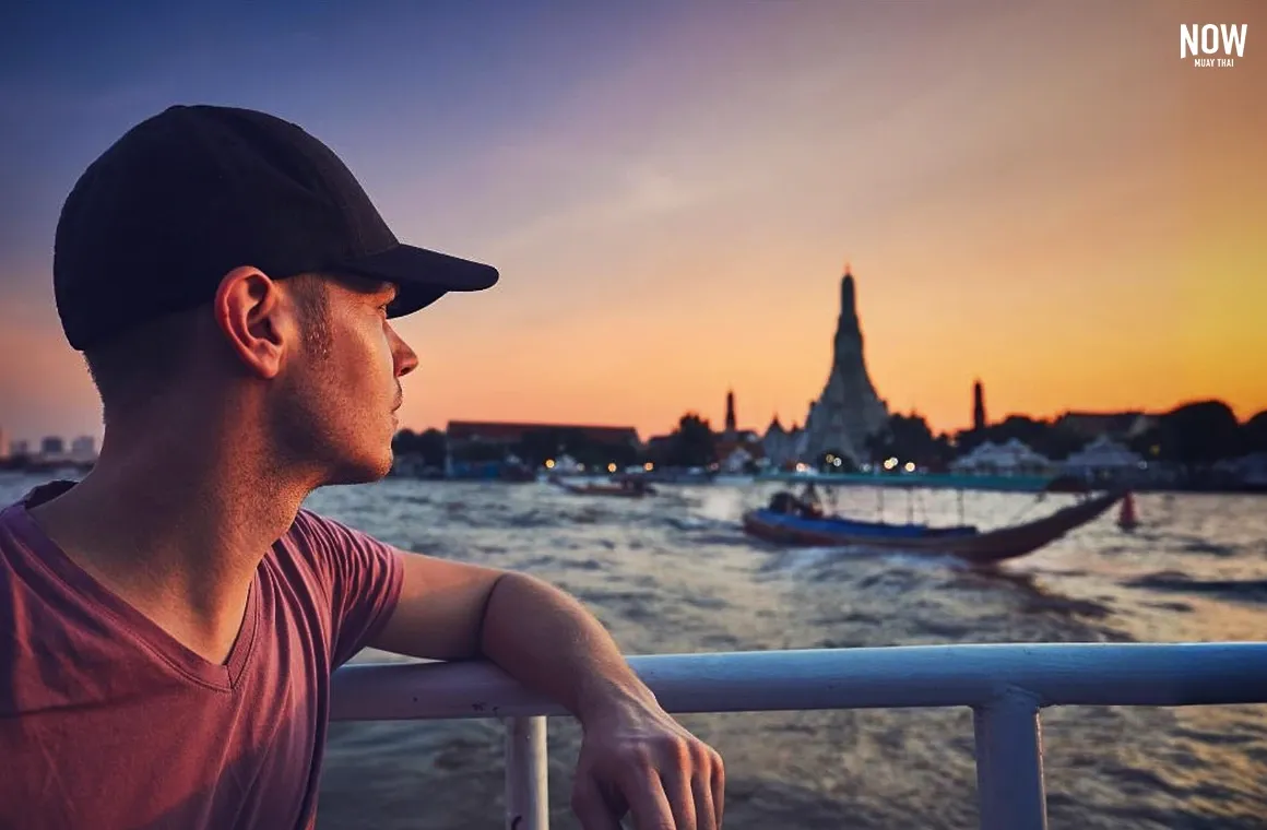 A man sits on a boat along the Chao Phraya River, with the stunning silhouette of Wat Arun illuminated by a golden sunset in the background.