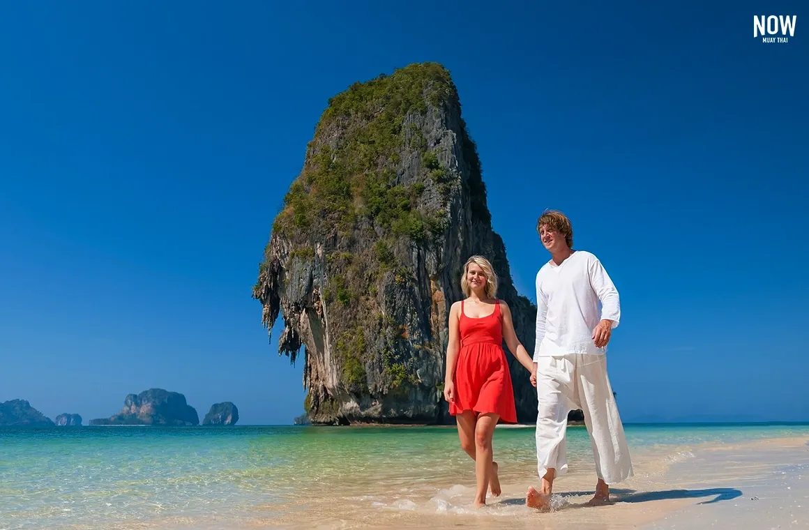 A happy family enjoys a summer vacation on the sandy beach of Koh Poda, Krabi, with a small sail-like islet rising prominently in the background, creating a picturesque scene.