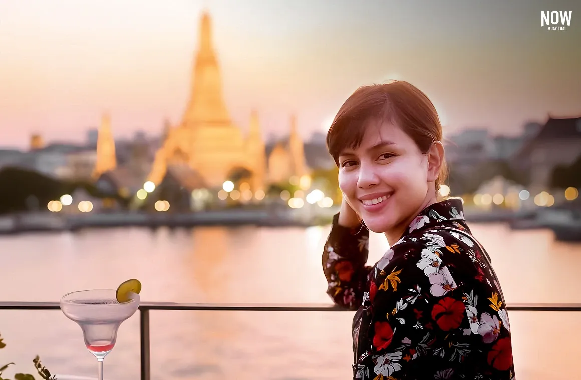 A woman tourist enjoys a cocktail while overlooking Bangkok's cityscape, with the iconic Wat Arun temple beautifully illuminated in the background.