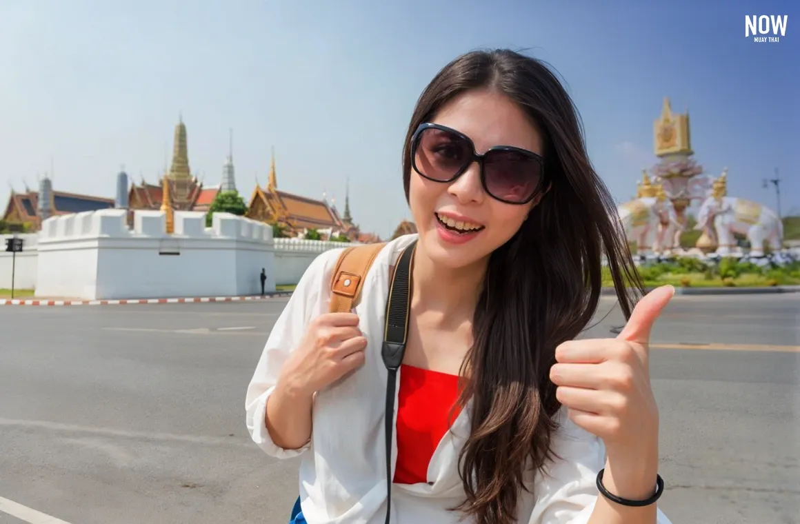 A happy female backpacker poses for a photo with the Emerald Buddha Temple (Wat Phra Kaew) and the Grand Palace in Bangkok, giving a thumbs-up gesture while exploring Thailand’s iconic landmarks.