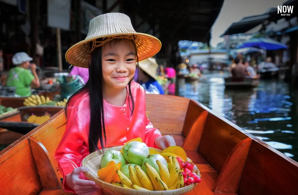 A young girl dressed in traditional Thai merchant attire poses for a photo while holding a tray of fruit, sitting on a boat at a floating market, symbolizing the traditional Thai culture where boats were commonly used for transportation in the past.