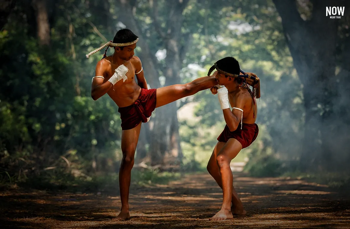 In the image, a Muay Thai fighter wearing traditional Muay Kard Chuek is engaged in ancient combat, with a Mongkhon on the head and Pra Jiad arm bands, performing the Wai Kru ritual before the fight.